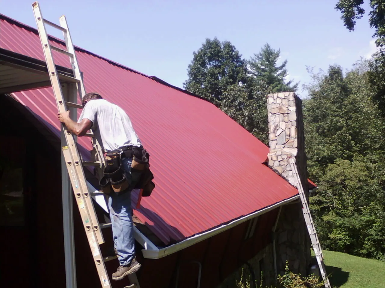 Roofer on a ladder beside a red metal roof and stone chimney — Soffit & Fascia Repair by Roofing Brothers in Big Bear City
